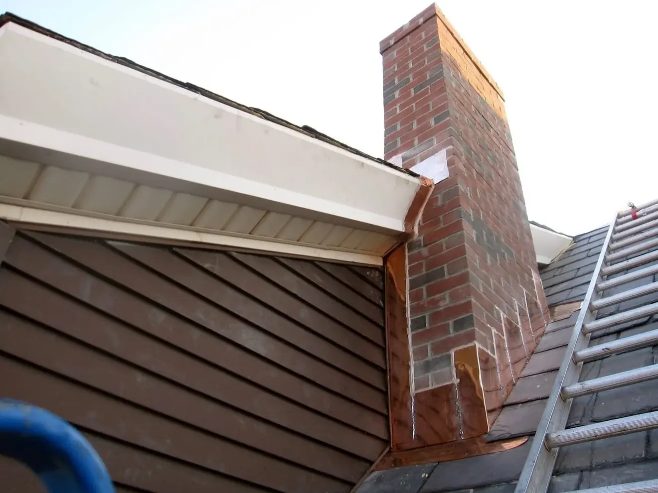 Copper flashing at a brick chimney on a slate roof, with white fascia and soffit along the roof edge