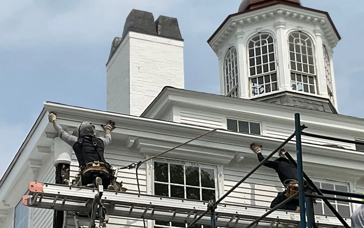 An Art Slate crew in harnesses working at the cornice of a historic white building from ladder staging, with a copper domed cupola behind them