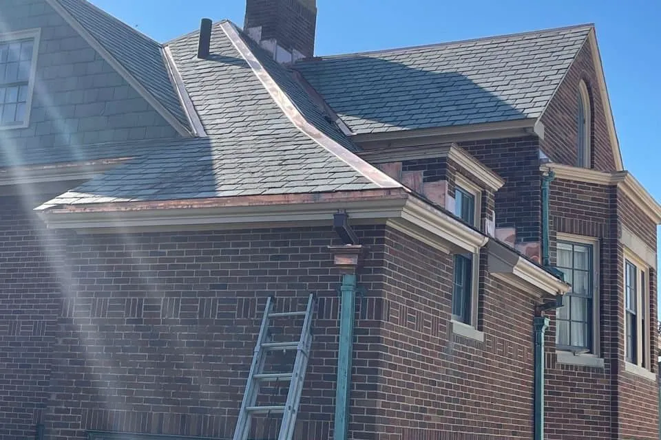 Early-1900s brick house with a new slate roof, a copper gutter and green patinated downspouts, and a ladder leaning on the brick wall below the eave, on an Art Slate project in Greater Boston