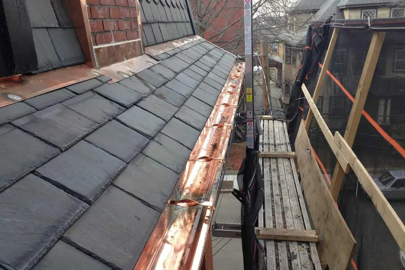 New copper gutter on hidden hangers along a slate roof edge next to pump jack staging and debris netting