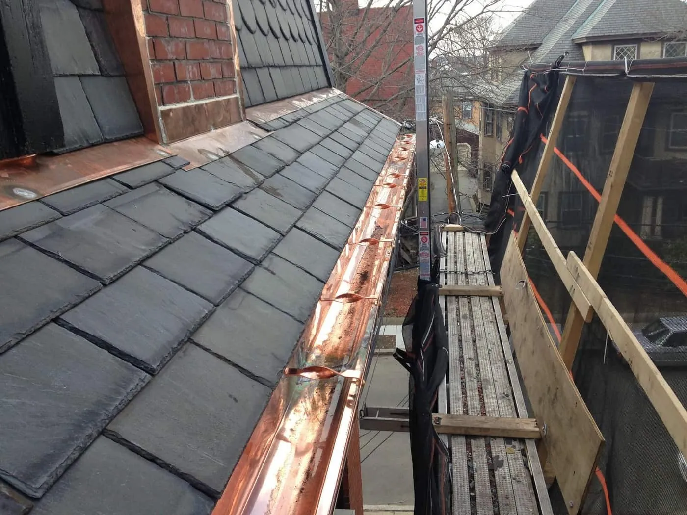 New copper gutter on hidden hangers along a slate roof edge beside pump jack staging and debris netting
