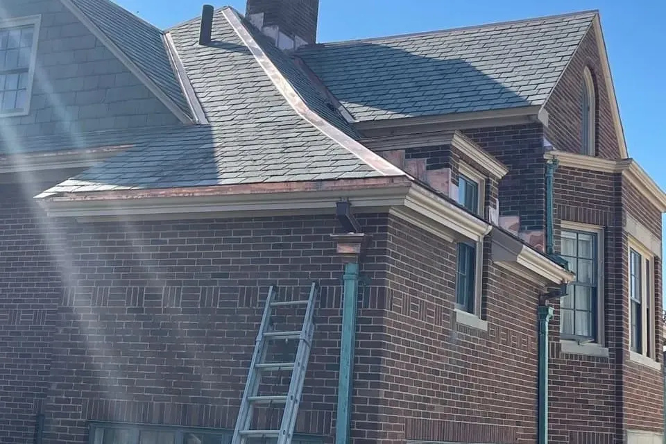 Brick house with a slate roof, copper gutters and green patina copper downspouts, with an extension ladder against the wall