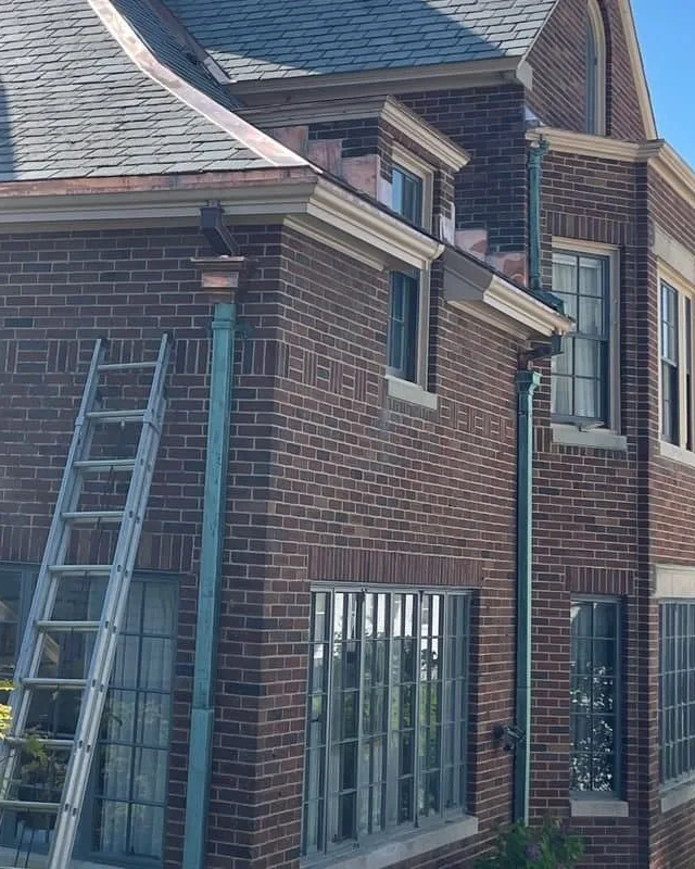 Copper leader heads and green patinated copper downspouts on an early-1900s brick house with a new slate roof, with a ladder against the brick, on an Art Slate project in Greater Boston