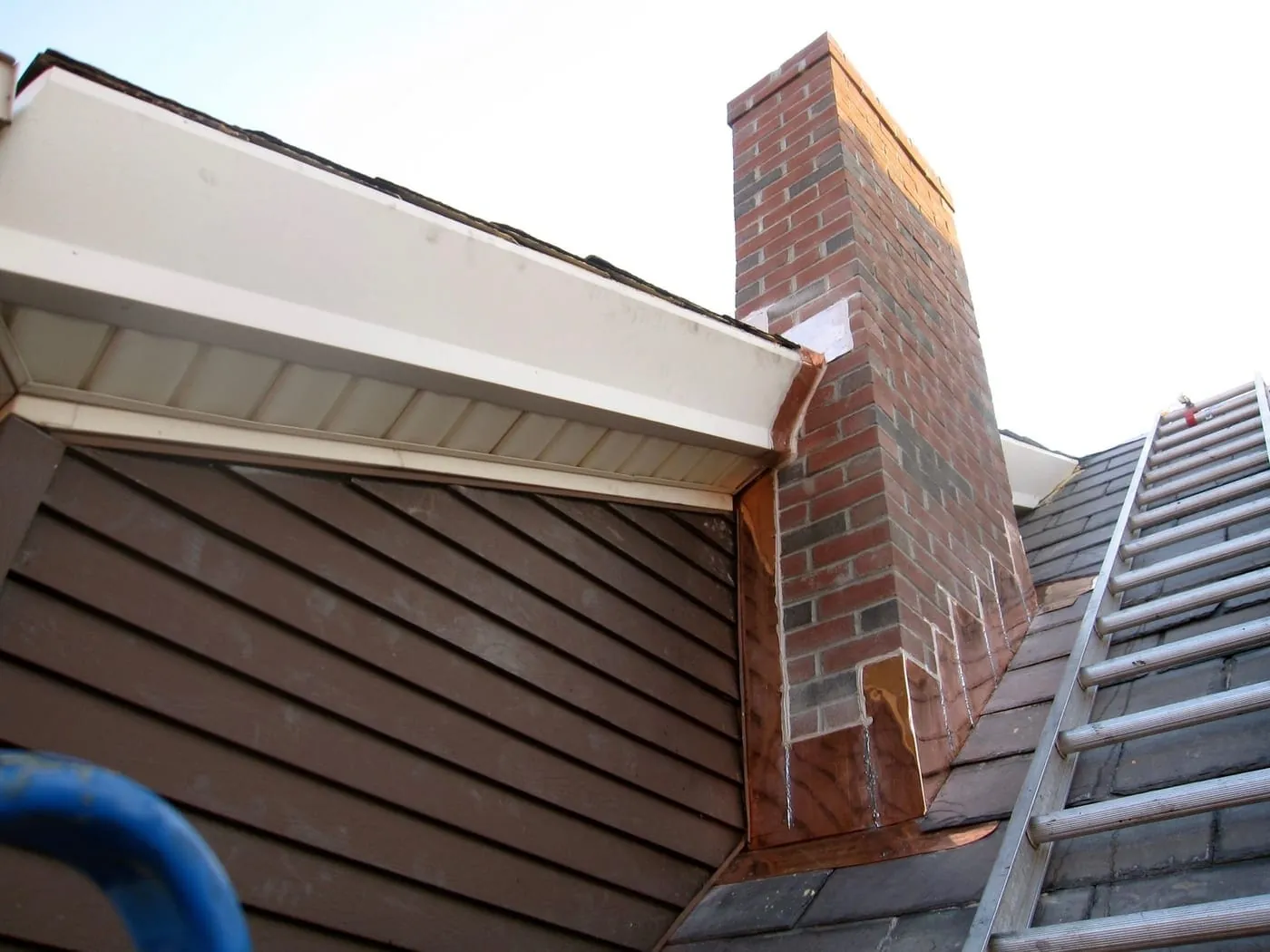 Brick chimney with copper step flashing on a slate roof, a ladder on the slate, and a white fascia and soffit at the roof edge