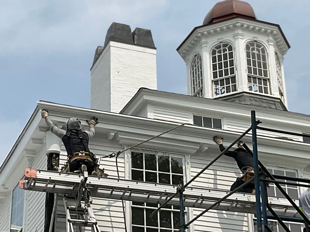 A crew in harnesses working at the cornice of a historic white clapboard building from ladder jack staging